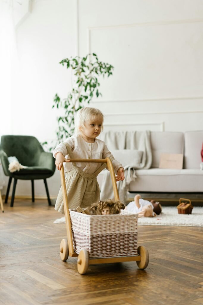 A young child pushes a wooden toy trolley filled with dolls across a living room floor, highlighting playful learning and safe indoor trolley use for children.