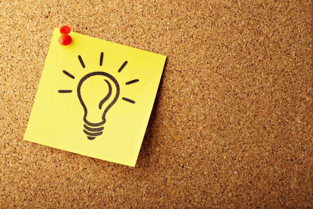 Yellow sticky note with hand-drawn lightbulb pinned to a corkboard, symbolising the spark of preliminary ideas and innovative thinking for future success in 2025