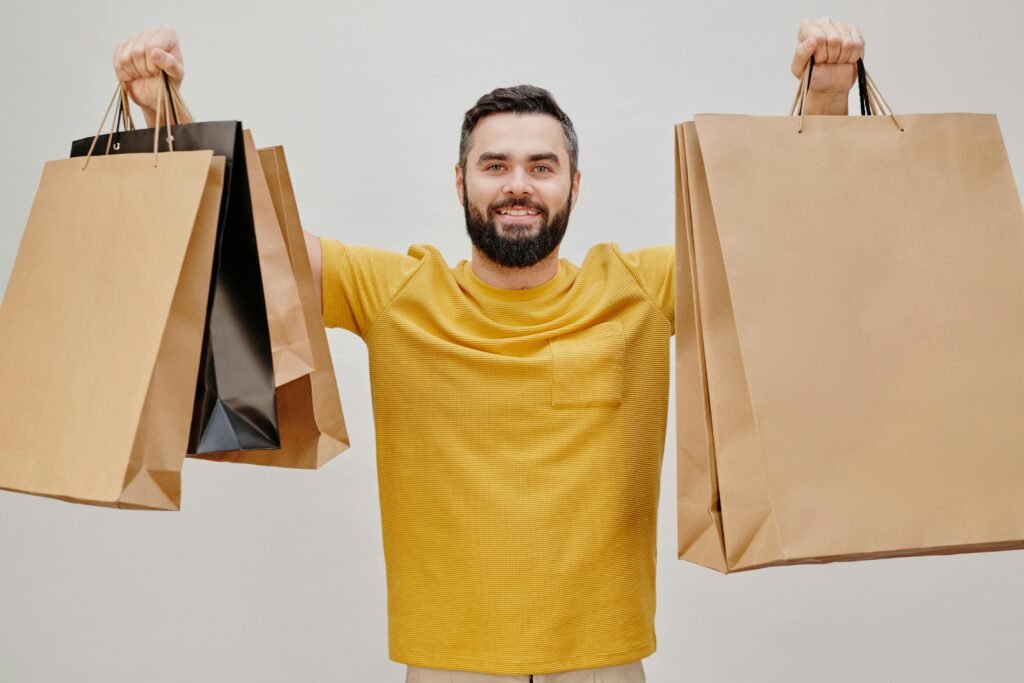A happy man in a yellow shirt holds up multiple large shopping bags, symbolising a rewarding retail experience—perfectly capturing the excitement and vibrant shopping environment of American Eagle’s Sahara Centre Sharjah 2025 expansion and the transformative style journey discussed in the article.